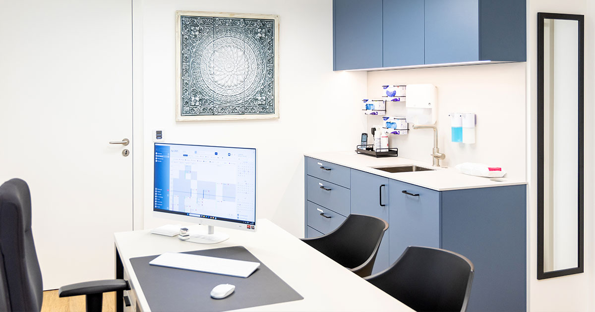 Modernes Behandlungszimmer im ROC Orthopädie Aschheim in München mit Schreibtisch, Computer, blauen Schränken und medizinischer Ausstattung.