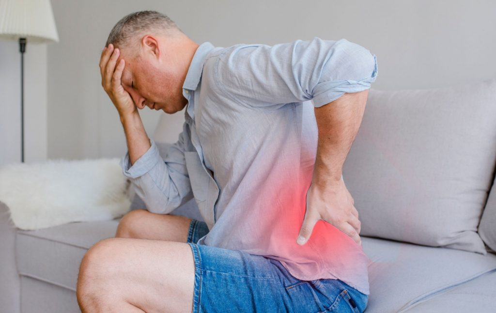 A man sits on the sofa with a pained expression on his face, holding his lower back—Racz catheter treatment for back pain at the ROC Regenerative Center in Aschheim