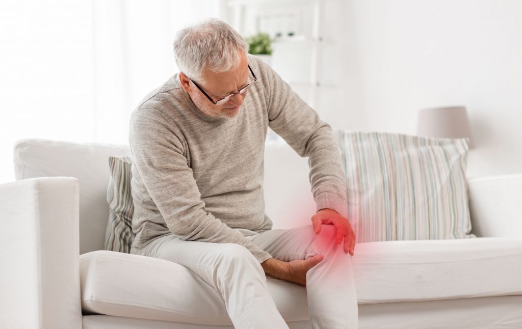 An elderly man sits on the sofa, writhing in pain, and holds his aching knee—stem cell therapy for knee pain at the ROC Regenerative Center in Aschheim
